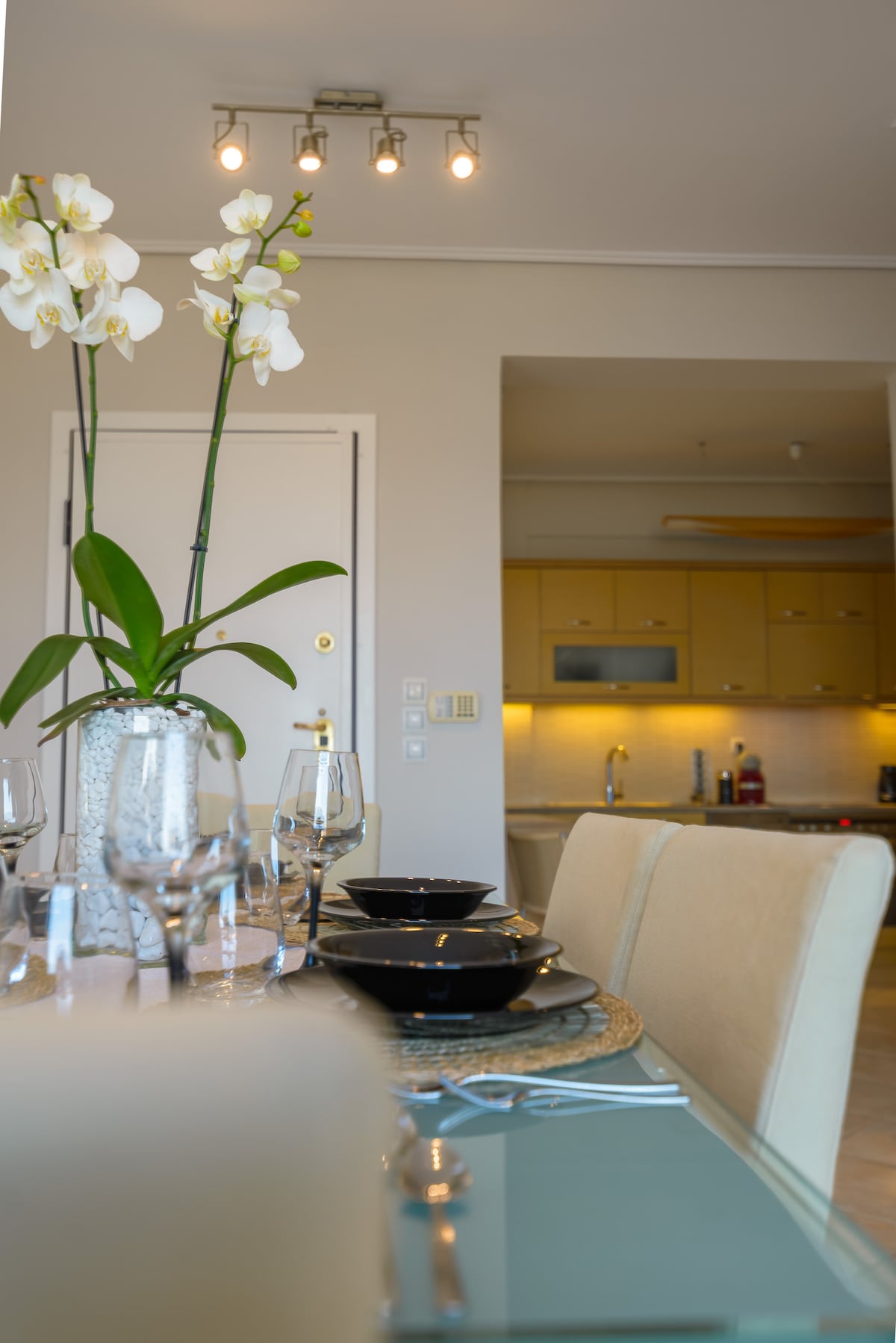 A dining area is set with an elegant glass table adorned with black dishware and sparkling glassware. A centerpiece of white orchids adds a touch of nature. In the background, a modern kitchen with yellow cabinetry can be seen, enhancing the space's warmth.