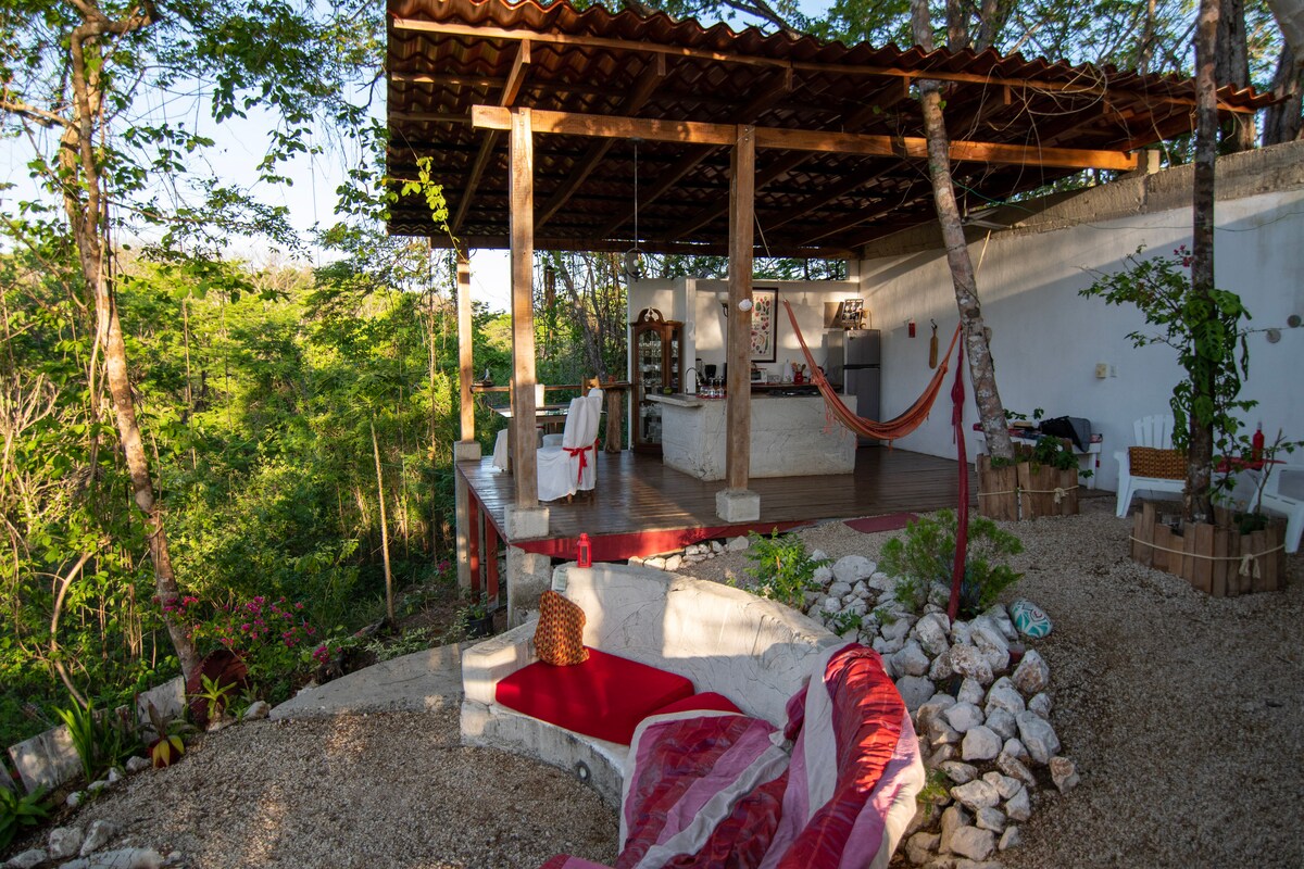 An open-air common area is viewed, featuring a hammock and seating arrangement on a wooden deck. Surrounding greenery enhances the tranquil setting, while the kitchen, partially visible, faces the jungle. A stone pathway leads to colorful floral accents, creating a seamless connection with nature.