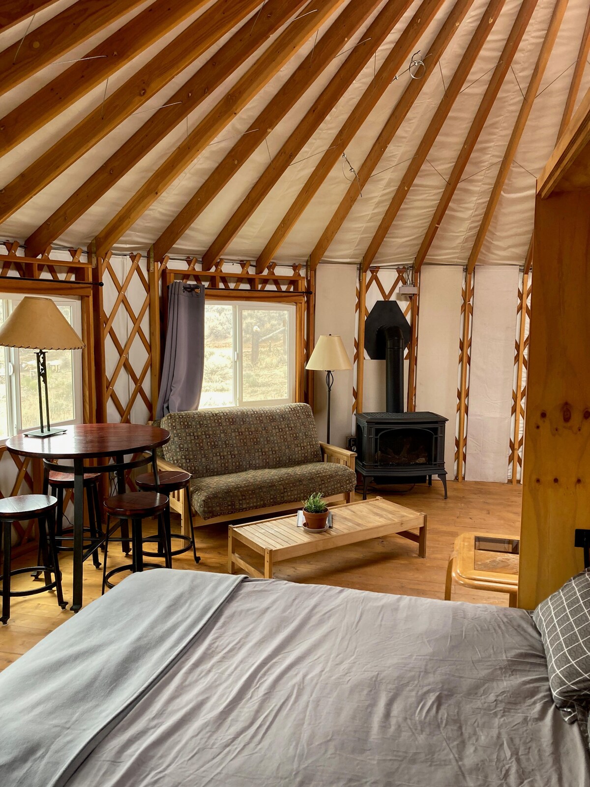 A spacious yurt interior showcases natural wood beams and bright windows, allowing natural light to fill the space. A comfortable couch and a wooden coffee table are accompanied by a small dining table with stools. A cozy wood stove adds warmth to the living area.