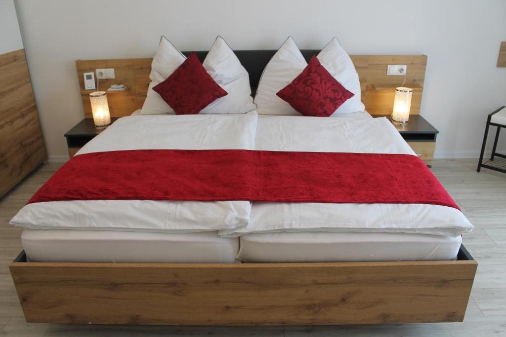 A spacious bed is presented with crisp white linens and decorative red pillows. The headboard features wooden panels on either side, accompanied by bedside lamps. Neutral-toned flooring complements the overall design, creating a calm ambiance.