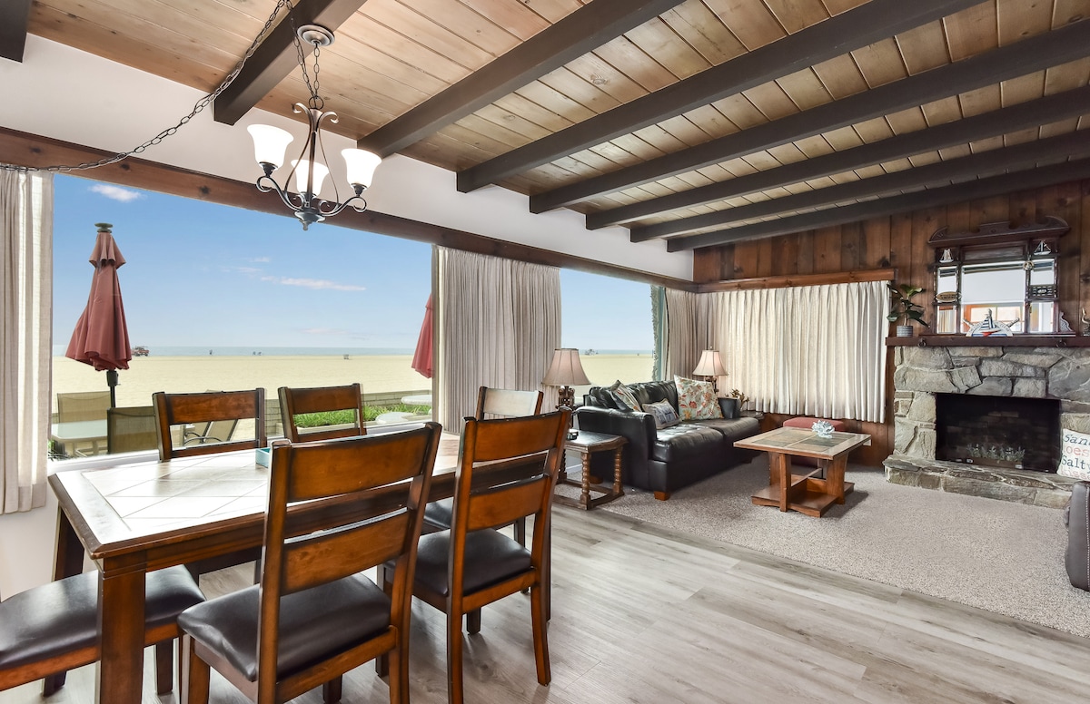 A spacious living and dining area features wooden beams overhead and large windows with ocean views. A rustic stone fireplace is present, along with leather couches and a dining table with wooden chairs. Natural light fills the space through sheer curtains.