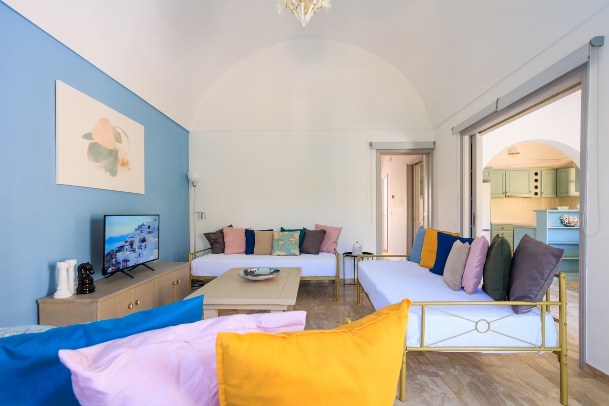 A bright and airy living space showcases a curved ceiling with soft lighting. Comfortable, multi-colored seating is arranged around a central coffee table. A television is positioned nearby, with views of a kitchen area visible in the background through open doors.