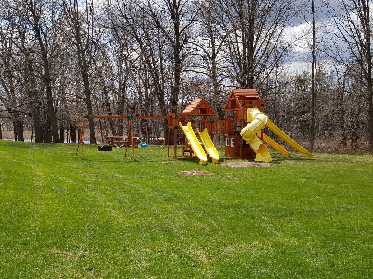 A wooden playset is situated on a grassy area, featuring two slides, a swing set, and a climbing structure. Surrounding trees provide partial shade, enhancing the outdoor play space. The well-maintained lawn complements the inviting atmosphere for children to enjoy.