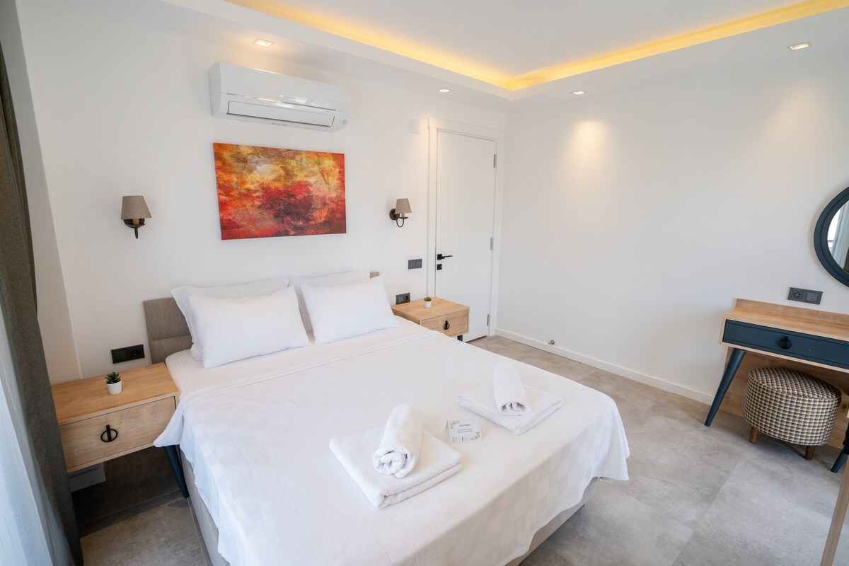 A spacious bedroom is presented with a large bed featuring neatly arranged towels. Light walls reflect natural light, complemented by wooden bedside tables. A vibrant artwork adds color to the space, while a mirror with a rounded frame and a woven basket provide additional design elements.