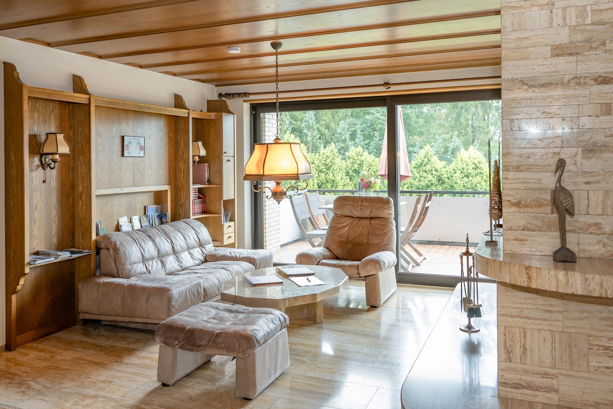 A spacious living area is featured, with light-colored leather seating arranged around a circular coffee table. Wooden shelves and a warm lamp create a cozy atmosphere, while large glass doors open to a balcony overlooking greenery.