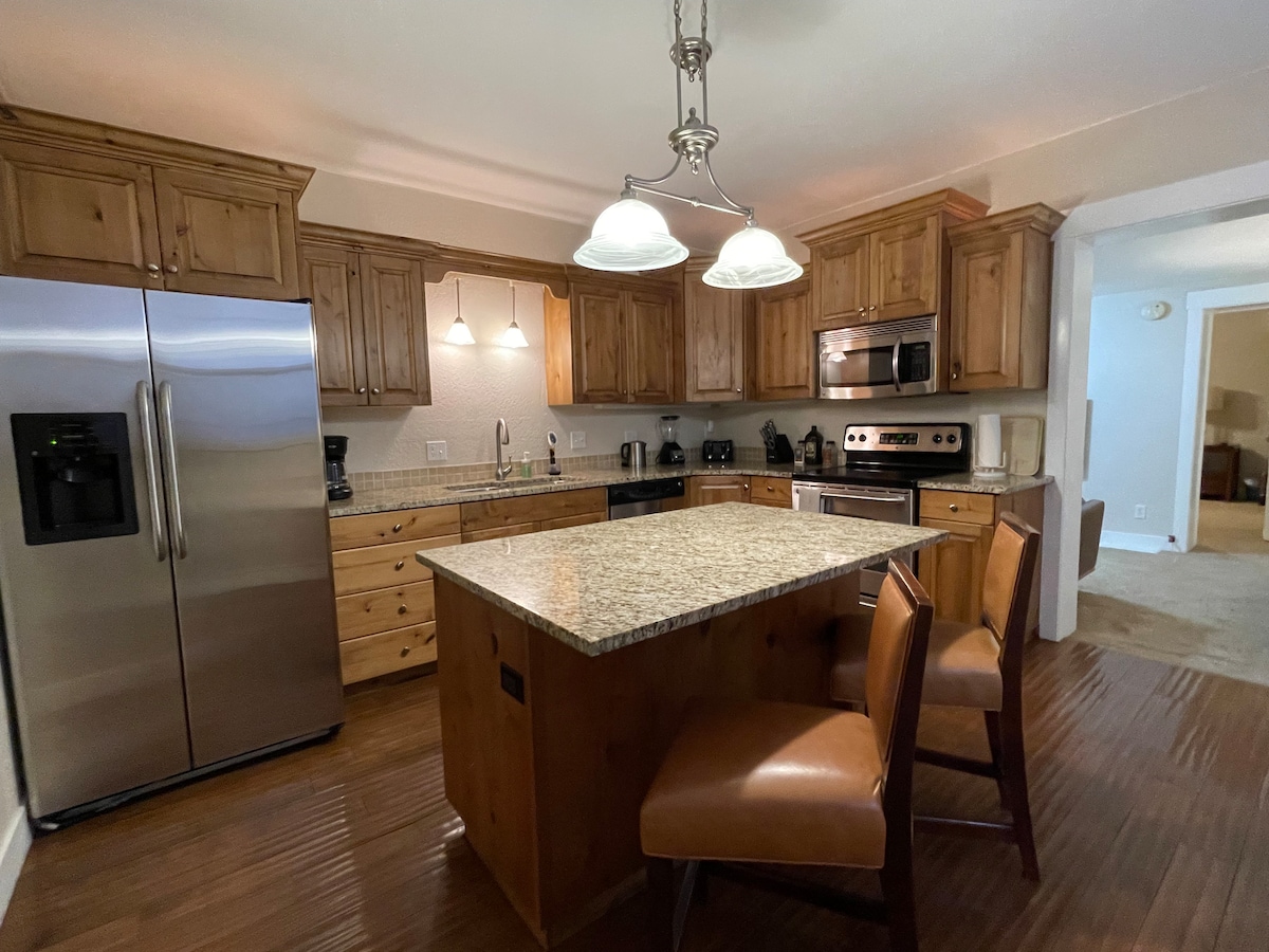 The kitchen features wooden cabinetry and a large island with two high-backed chairs. Stainless steel appliances, including a refrigerator and oven, complement the granite countertops. Warm lighting from pendant fixtures enhances the inviting space, while the open layout connects to adjacent areas.