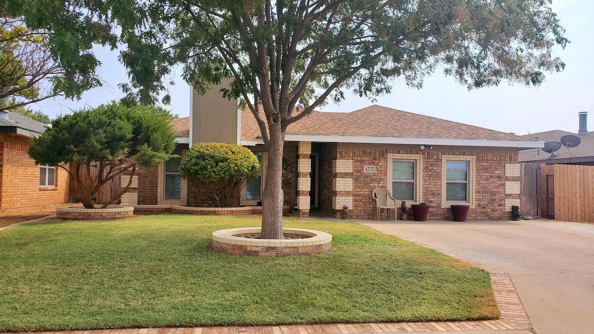 The exterior of a single-story brick home is featured, framed by a well-maintained lawn and a large tree. Planters with lush greenery are visible near the entrance, alongside two maroon chairs on a paved driveway leading up to the house.
