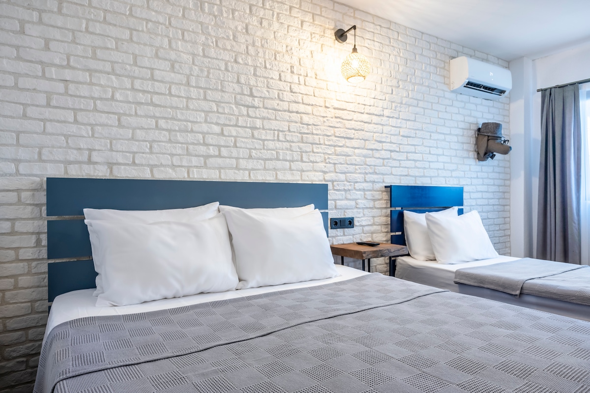 A sleeping area features two spacious beds with white linens, positioned against a textured light-colored brick wall. Soft lighting is provided by a hanging fixture above the beds. A small wooden table is placed beside one bed, enhancing the room's functionality.