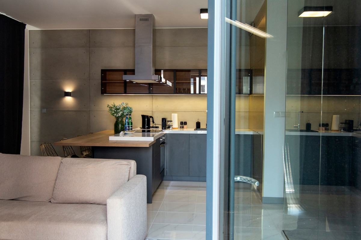 A modern kitchen area with sleek cabinetry and a central island is featured. Stainless steel appliances include a hood and coffee maker. A light-filled living space with a comfortable sofa offers a view through large glass doors that extend to an outdoor area.