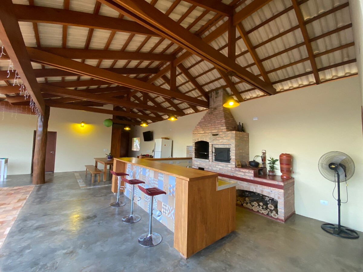 An open and airy kitchen space is highlighted by a wooden bar with seating. A rustic stone fireplace is central to the room, framed by high wooden beams and warm lighting fixtures. A fan is positioned in the corner for comfort.