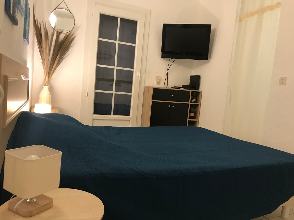 A comfortable sleeping area features a spacious bed covered in a deep blue quilt. A wall-mounted TV is visible above a simple cabinet. Soft lighting is provided by a bedside lamp, while a large window with a frosted curtain allows natural light to enter.
