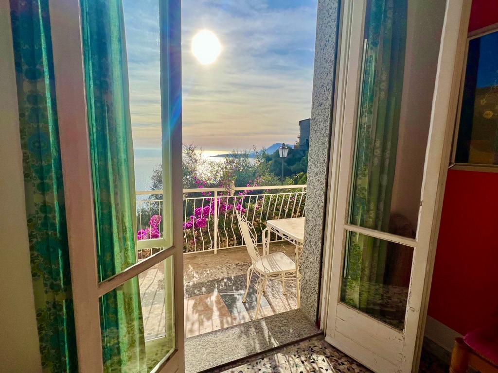 Natural light fills the space as double doors open to a private balcony adorned with blooming bougainvillea. A small table and chairs are set for outdoor enjoyment, while the tranquil view overlooks the sea and a vibrant sunset.