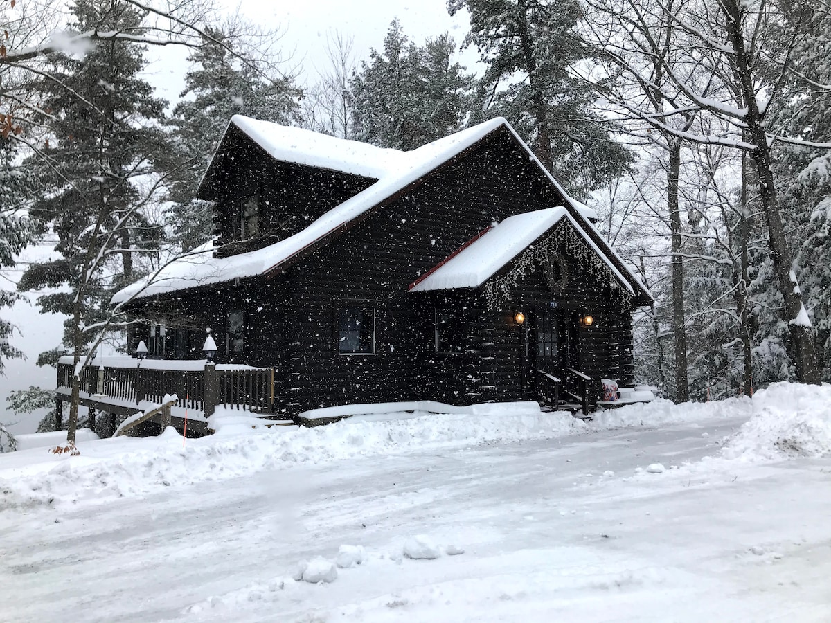 A log cabin stands in a serene winter landscape, blanketed in freshly fallen snow. Dark wooden walls contrast with the white surroundings, while the sloped roof sheds the snow. Snowflakes gently fall, creating a tranquil ambiance amid the surrounding trees.