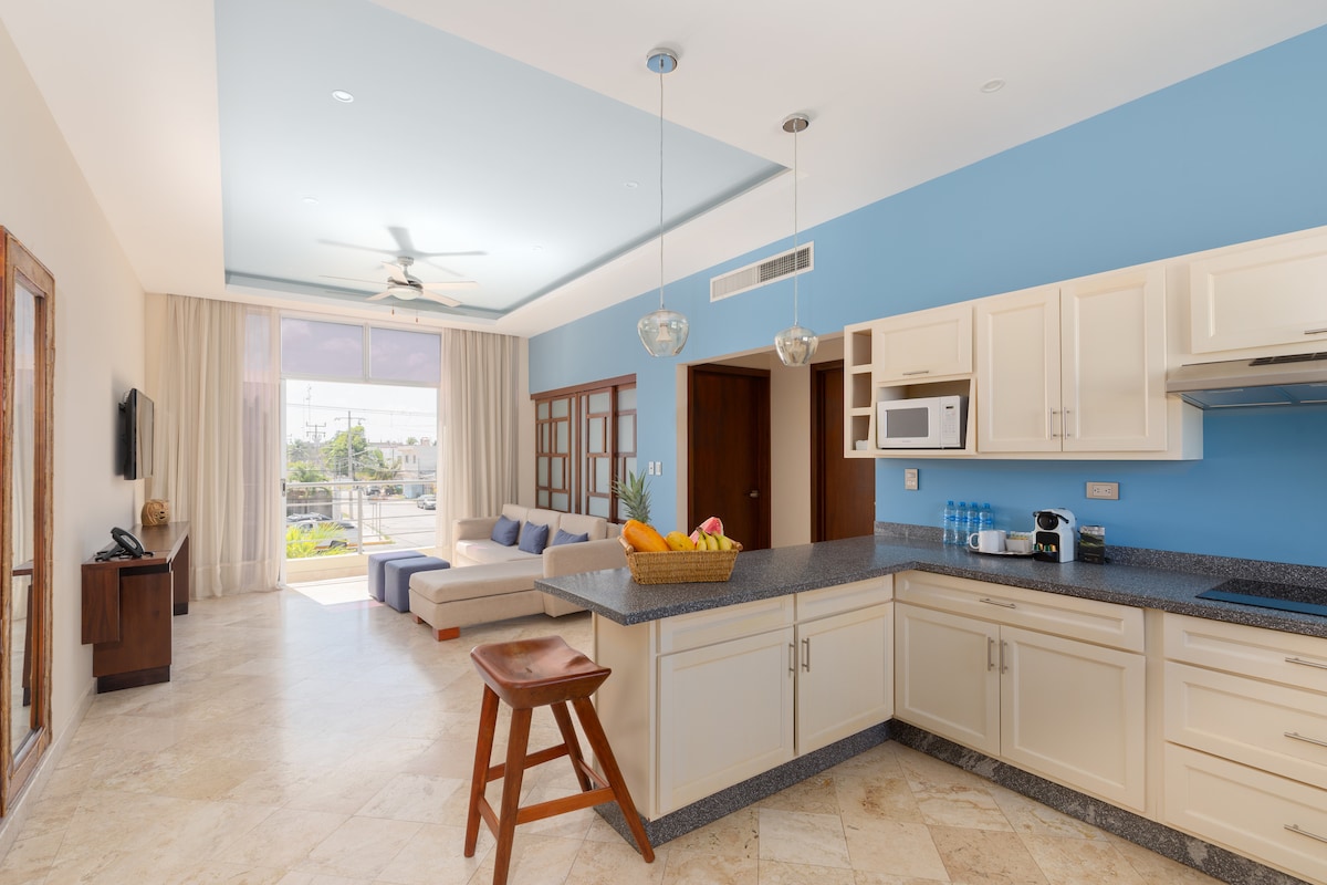 A modern kitchen features light cabinetry and a granite countertop, with a spacious living area visible. A large window brings natural light into the open space, where a comfortable seating area and a fruit basket are displayed. Light blue and natural tones create a relaxed atmosphere.