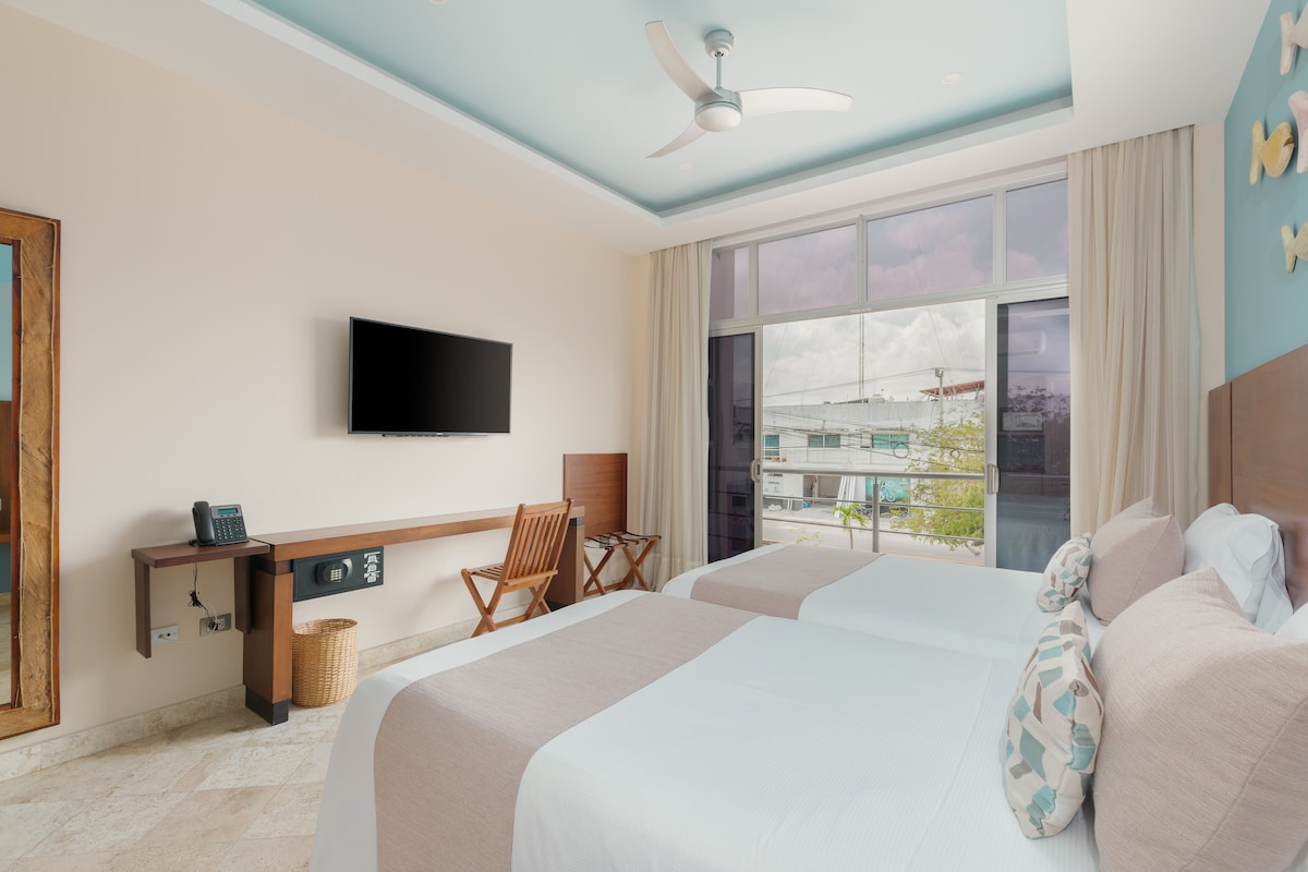 A light-filled room features two individual beds positioned against a wall. A flat-screen television is mounted above a wooden console, while a vintage-style folding chair is placed nearby. Large windows offer views to the city, adorned with light curtains.