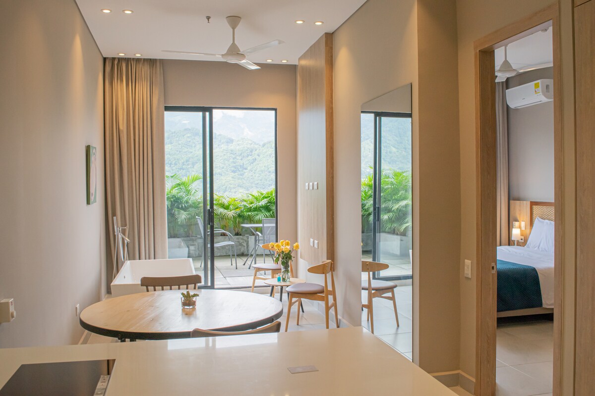 A spacious interior is captured with an open layout. A circular dining table is surrounded by two wooden chairs, while a light color palette enhances the ambiance. Large glass doors lead to a terrace adorned with greenery, offering views of the surrounding mountains.