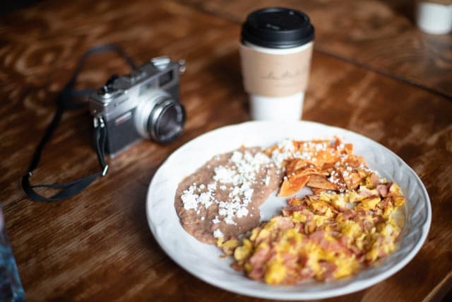 A breakfast plate is presented on a textured wooden table, featuring scrambled eggs, tortillas, and beans topped with cheese. A coffee cup and a camera are positioned nearby, adding a casual touch to the setting.