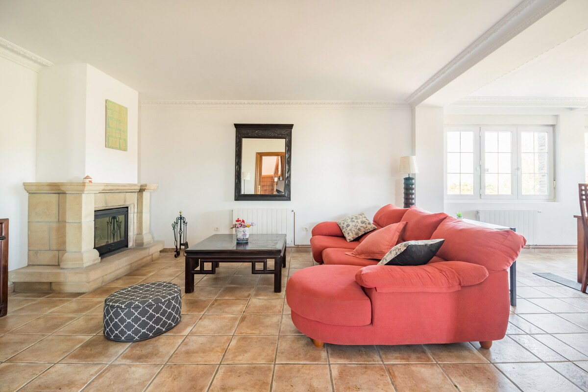 A spacious living area features a red sectional sofa and a low wooden coffee table. Light enters through multiple windows, illuminating the stone fireplace and creating a bright atmosphere. A decorative mirror adds depth, complementing the neutral tones of the tile flooring.