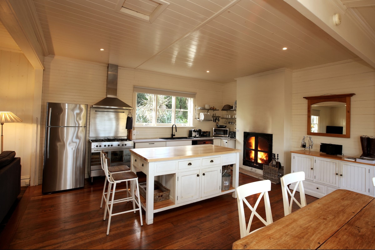 A spacious kitchen is depicted, featuring a large island with a wooden countertop and seating for four. Stainless steel appliances are visible, including a refrigerator and a gas stove. A cozy open fireplace adds warmth to the room, with natural light flowing through a wide window.