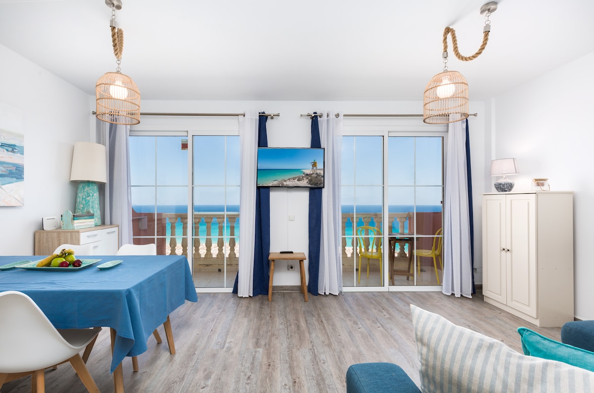 A bright living area features large glass doors leading to a balcony with ocean views. A wooden dining table is set with a blue cloth, while light-colored furniture and decor create a relaxed setting. Two pendant lights hang from the ceiling, enhancing the spacious feel.