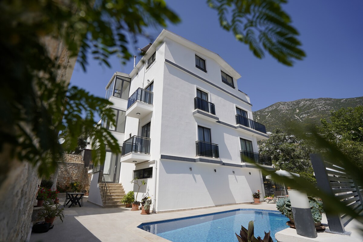 A modern three-story building is showcased, featuring multiple balconies and large windows. The entrance is framed by lush greenery, and a clear pool can be seen reflecting the blue sky. The surrounding area includes potted plants adding a touch of nature.