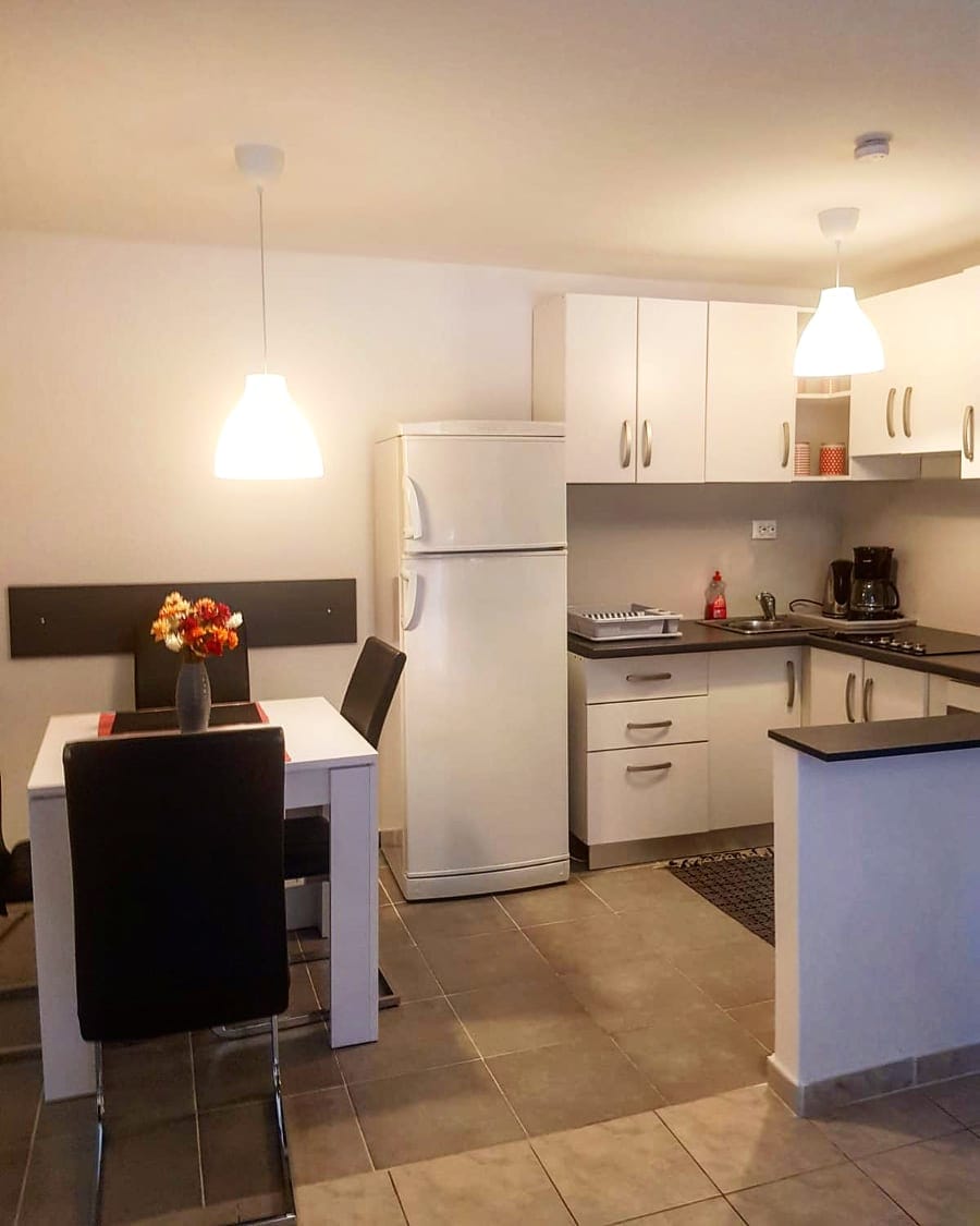 A functional kitchen area is displayed, featuring white cabinets and a sleek black countertop. A dining table with four black chairs is set beneath soft lighting fixtures. A refrigerator, stove, and coffee maker are visibly arranged, contributing to the practicality of the space.