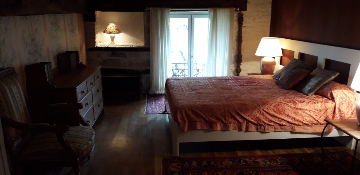 A cozy private room features a double bed with an ornate coverlet, complemented by bedside lamps. A wooden dresser and a vintage armchair are present, along with light-filtering curtains that frame a window overlooking the exterior. Natural wooden flooring enhances the room's charm.