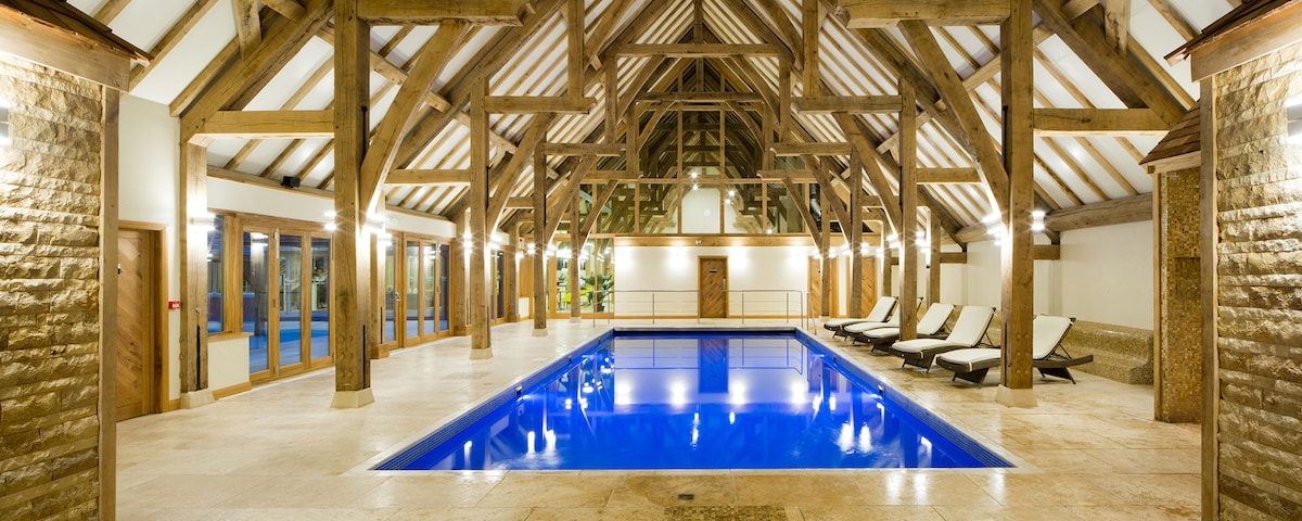 An indoor swimming pool is framed by wooden beams under a high ceiling. Sun loungers are positioned alongside the pool, which is filled with clear blue water. Large windows allow natural light to fill the spacious area, enhancing the elegant ambiance.