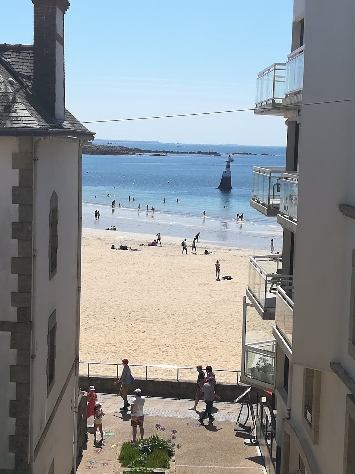 A view from the balcony showcases the sandy beach and calm waters. People are seen enjoying the sun and water, while a lighthouse is visible in the distance. The image captures the lively atmosphere of a beach day framed by nearby buildings.