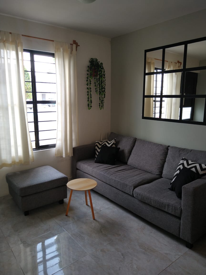 A cozy living area is furnished with a gray sofa and a matching ottoman. A small round wooden table is positioned nearby. Natural light enters through large windows, complemented by sheer curtains. A decorative mirror and a small plant add visual interest to the space.