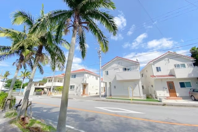 A row of modern villas is presented along a tree-lined street. Palm trees are noticeable in the foreground, while a clear blue sky with scattered clouds provides a bright backdrop. The villas feature light-colored exteriors, with red-tiled roofs and simple, functional designs.