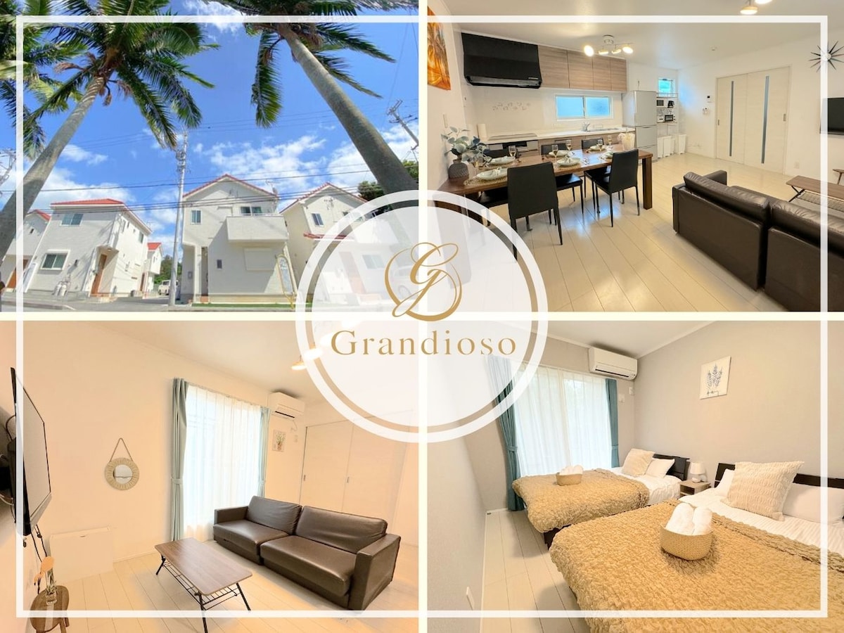 The image showcases a two-story villa surrounded by palm trees under a bright blue sky. The interior features a modern kitchen and dining area, along with a comfortable living room with a dark sofa. A cozy sleeping area is visible with beige bedding.