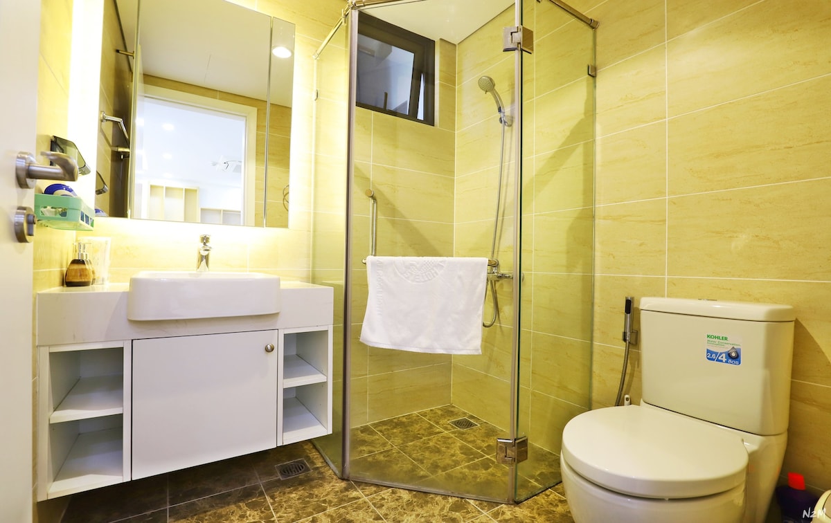 The modern bathroom features a glass shower enclosure and a spacious white sink atop a light wooden vanity. A towel hangs neatly for convenience, while a stylish toilet is positioned next to the vanity. Neutral tones and sleek finishes contribute to a clean and functional space.