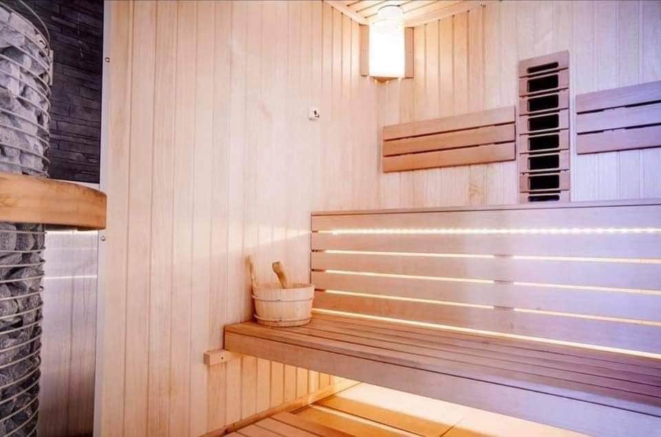 A sauna is depicted with warm wooden paneling and benches. Soft lighting is provided by a ceiling fixture and illuminated strips along the benches. A wooden bucket is placed on the lower bench, while a stone heater stands against one wall, enhancing the serene environment.