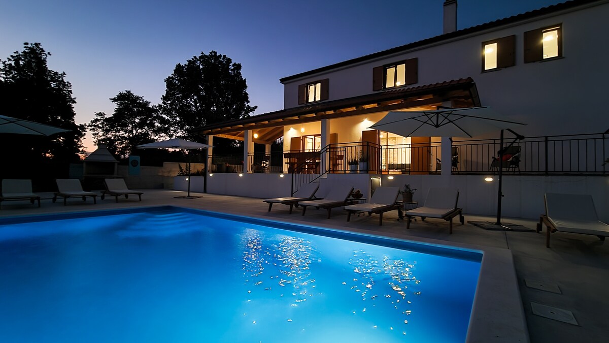 A serene outdoor space is highlighted, featuring a clear blue pool reflecting evening lights. The two-story house, with large windows, invites a warm ambiance. Comfortable lounge chairs are arranged around the pool, and soft shadows from nearby trees enhance the tranquil setting.