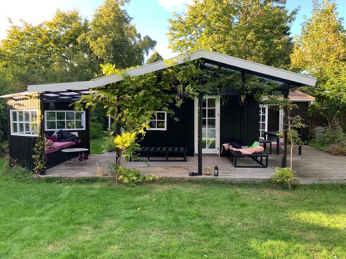 The exterior of a dark wooden summer house is surrounded by green grass and trees. A spacious wooden terrace is visible, featuring a simple outdoor seating area with cushions. Large windows add to the inviting atmosphere, allowing natural light to enhance the space.