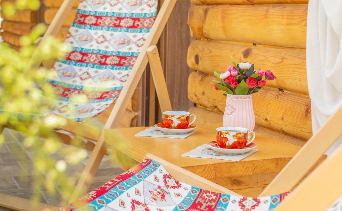 Two lightweight loungers are positioned beside a small wooden table. The table is set with two decorative teacups and a vase containing a bouquet of vibrant flowers. Natural wood walls are visible in the background, creating a harmonious outdoor lounging area.