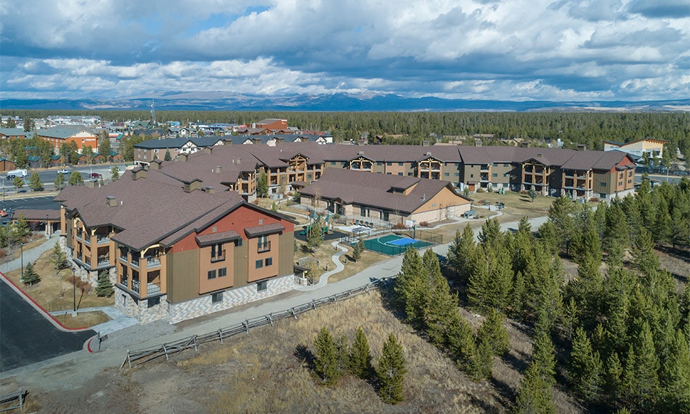 An aerial view captures the resort complex, showcasing a cluster of buildings with distinct architectural styles surrounded by trees and open landscape. Paths meander through the grounds, and amenities like a pool can be seen, all set against a backdrop of cloudy blue skies.