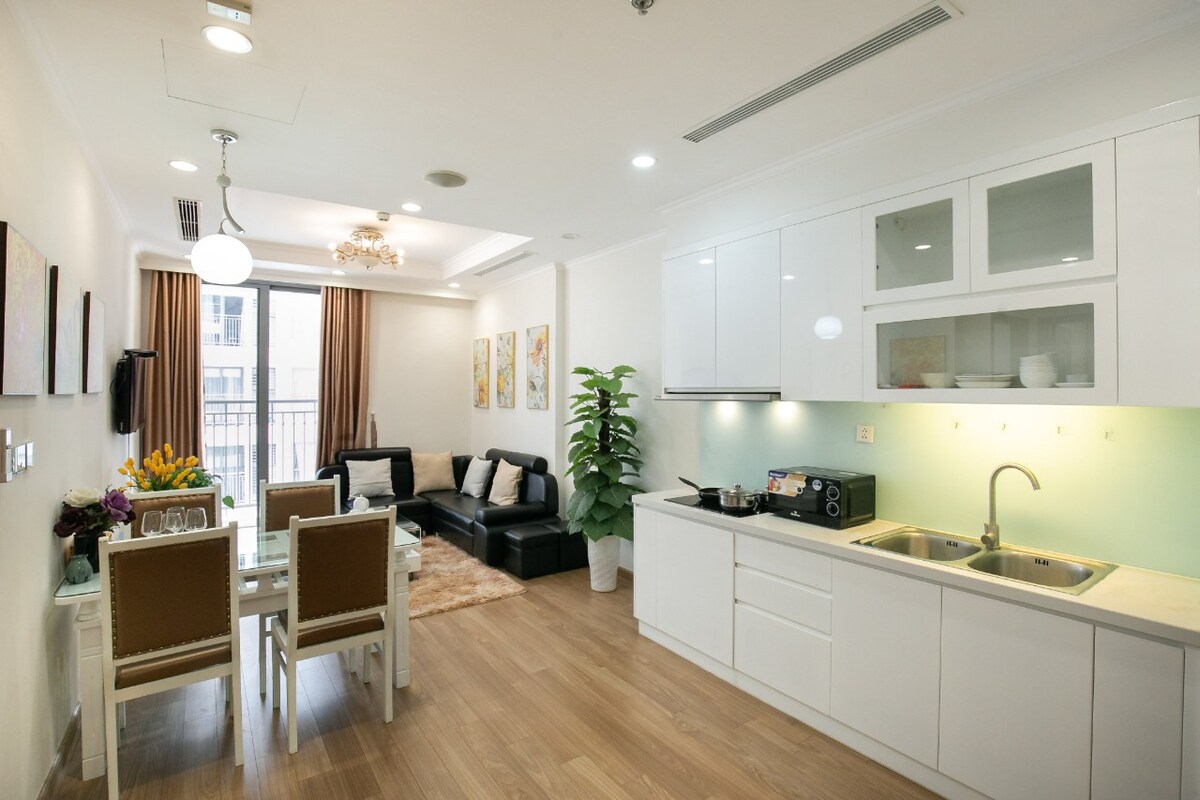 A modern open-plan living area features a stylish black sofa and a sleek dining table with four chairs. Bright artwork adorns the walls, complemented by natural light from large windows. The kitchen is equipped with contemporary cabinetry and essential appliances.