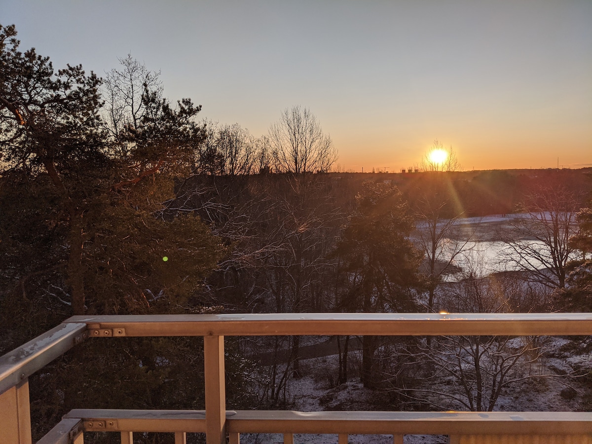 The image captures a scenic view from a spacious balcony at sunset. The warm glow of the sun illuminates the landscape, highlighting the trees and nearby water. A serene atmosphere is created by the natural surroundings, with a pathway gently winding through the area.