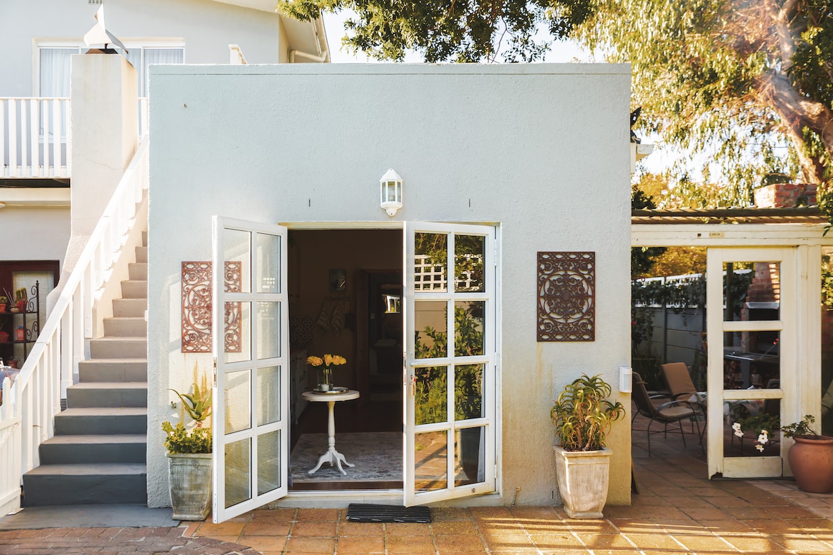 The exterior of a self-catering apartment is displayed, featuring double doors that open onto a welcoming entryway. A small round table is positioned on the outdoor patio, surrounded by potted plants. A staircase leads to the upper level of the adjoining property.