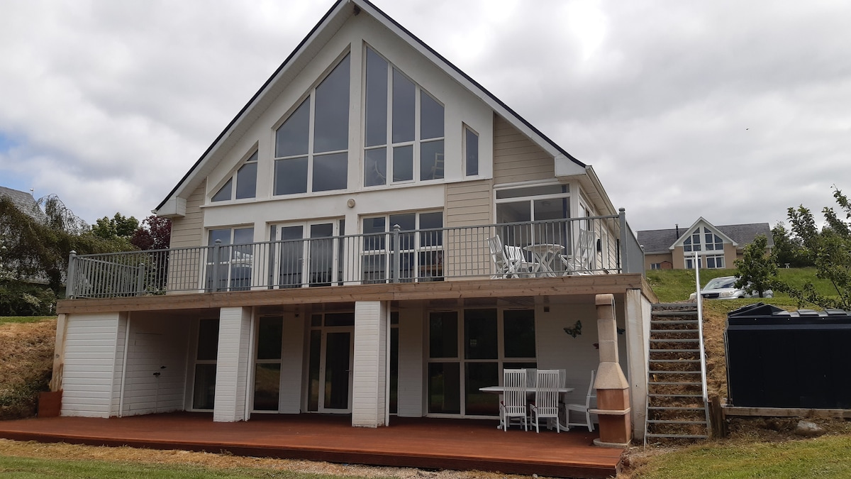 The exterior of a spacious house is presented, showcasing a large deck with outdoor seating and a dining table. Above, large windows flood the interior with light. A staircase leads to the upper level, while a grassy area can be seen in the background.