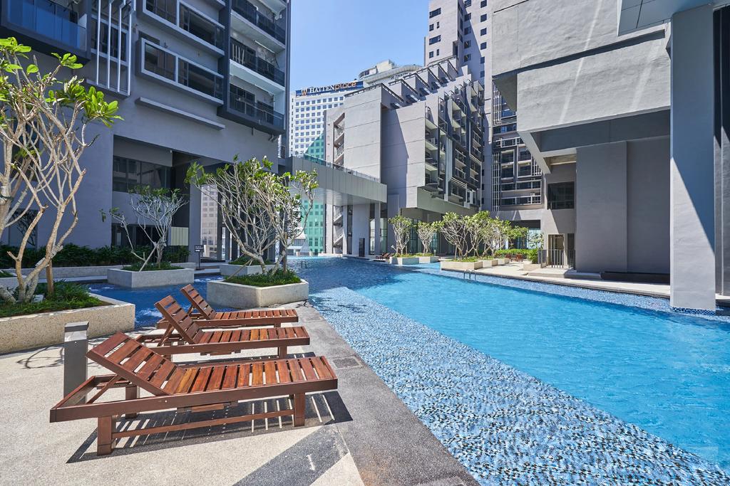 Lounge chairs are arranged along the edge of a clear blue pool, surrounded by modern architecture and greenery. The pool area features minimalist design elements and is bathed in natural light, creating a refreshing environment.