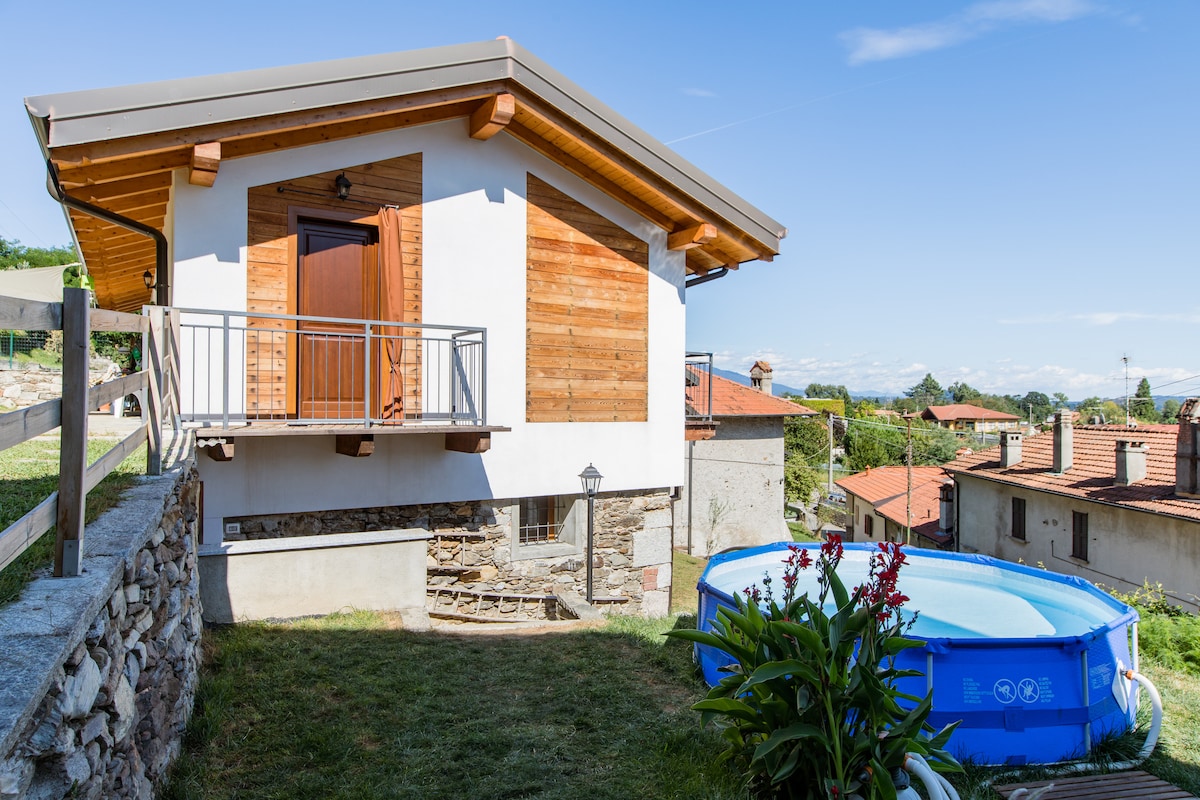 A charming villa exterior is displayed, featuring wooden accents and a covered entrance. A round above-ground pool is positioned on the grassy area, surrounded by plants and flowers. The serene countryside and nearby buildings are visible in the background under a clear blue sky.