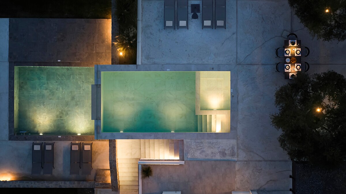 An aerial view reveals a private pool, surrounded by sunbeds and illuminated by soft lights. The patio includes a large dining table set for gatherings, contrasting with the serene ambiance of the surrounding olive groves.