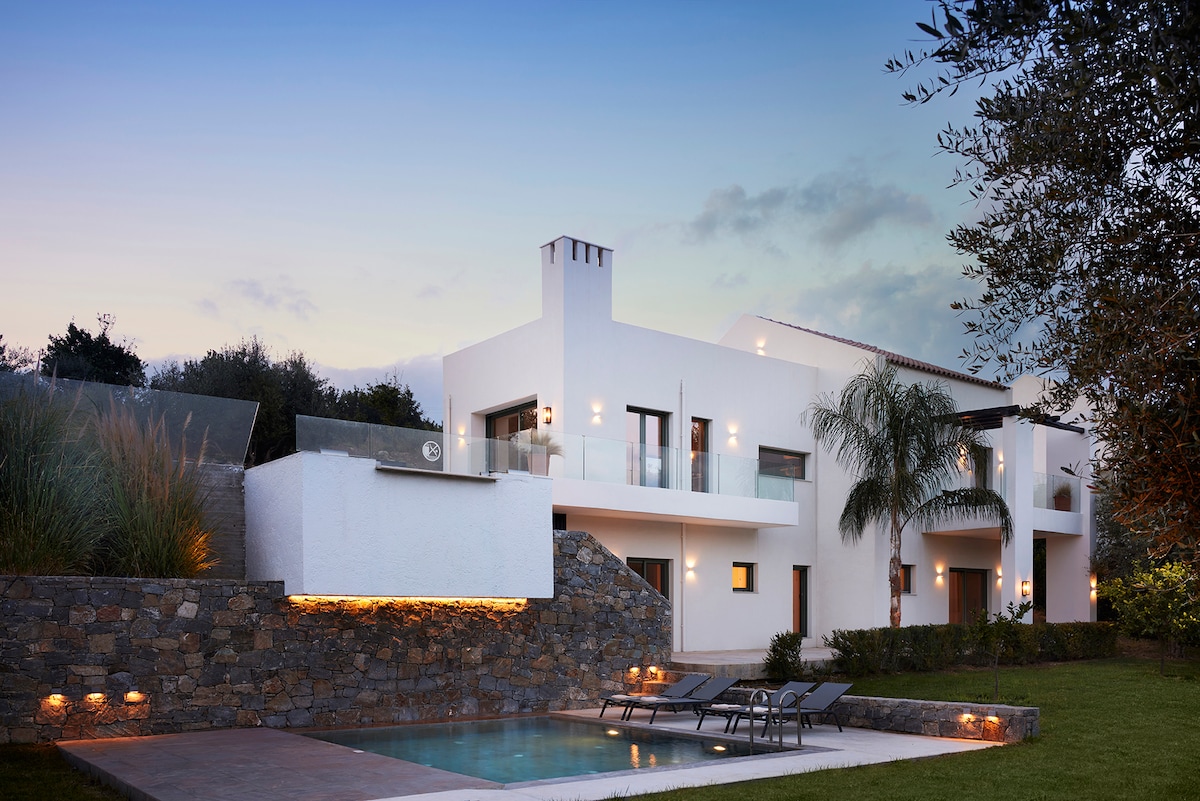The exterior of Agrielia Villa is showcased in a serene evening setting, featuring clean lines and modern architecture amidst olive trees. A private pool with comfortable sunbeds is highlighted in the foreground, while soft lighting enhances the tranquility of the space.