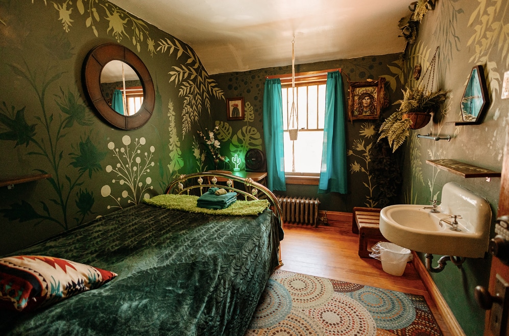 A cozy bedroom is adorned with leafy green wall decor, enhancing a serene atmosphere. A spacious bed is covered with a soft green blanket, while a small sink is positioned in one corner. Bright turquoise curtains frame a window, allowing natural light to brighten the space.
