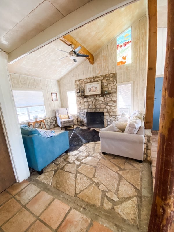 A spacious living area is highlighted by a stone fireplace and natural wood beams. Two comfortable sofas, one in light blue and one in white, are arranged around a textured stone floor. Large windows allow natural light to fill the room.