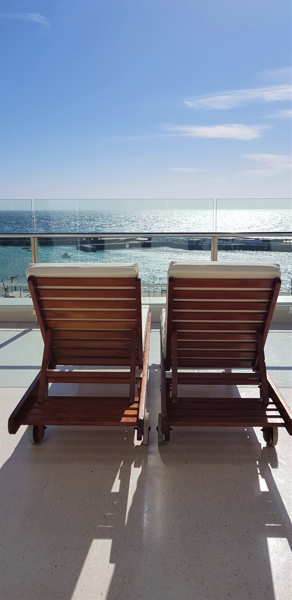 Two wooden lounge chairs are positioned in front of a glass railing, offering unobstructed views of the ocean. Sunlight reflects off the water, creating a bright and inviting atmosphere. The serene setting suggests a perfect spot for relaxation and enjoying the coastal scenery.