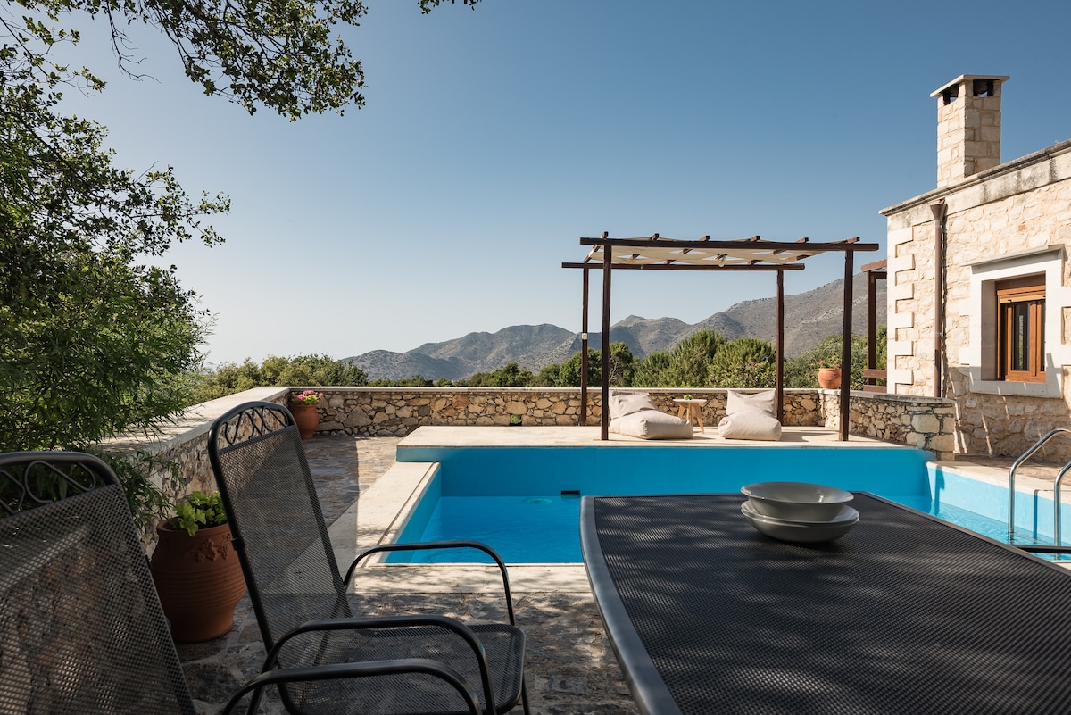 A serene outdoor area features a private pool surrounded by stone walls, complemented by shaded seating. A dining table is set with dishes, offering a view of the distant mountains. Potted plants add a touch of greenery to the inviting setting.